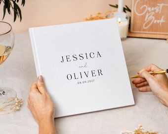 May include: A square white guest book with a matte finish featuring black printed text that reads JESSICA and OLIVER followed by the date 08.09.2027. The cover has a clean, minimalist design with centered typography in a serif font style.