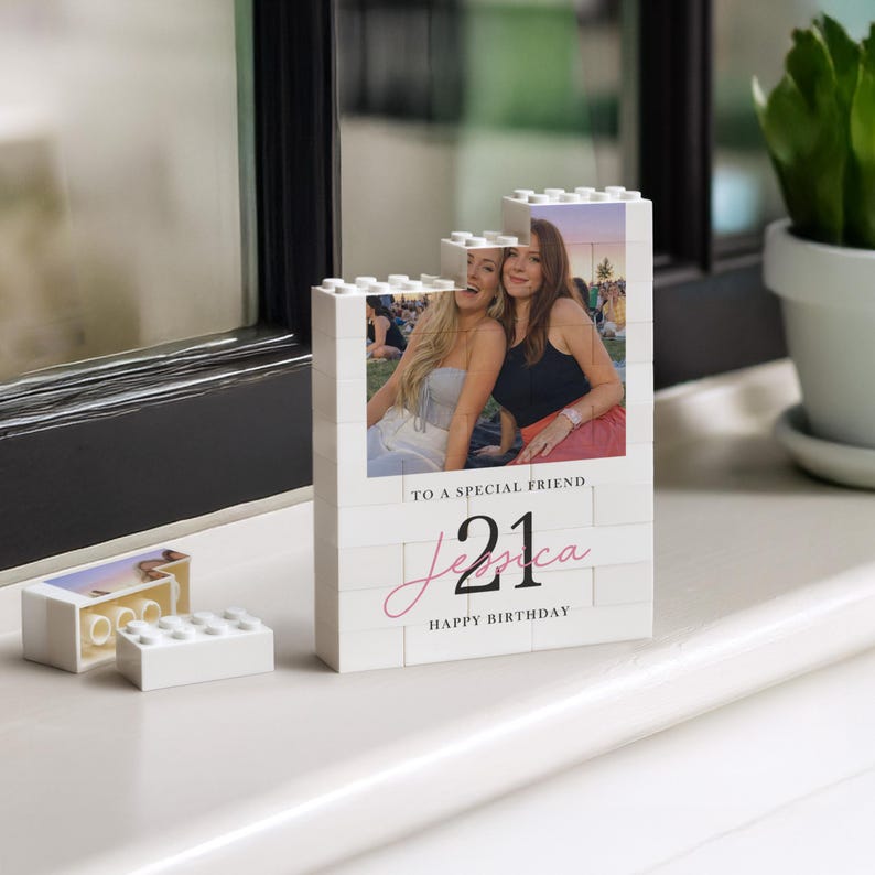 A white photo frame with a photo of two smiling women, placed on a white surface next to a potted plant.