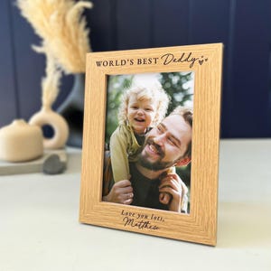 May include: A light oak wood picture frame featuring the engraved text WORLD'S BEST Daddy with two small hearts at the top and Love you lots, Matthew at the bottom. The frame holds a photograph of a smiling child being held by an adult.
