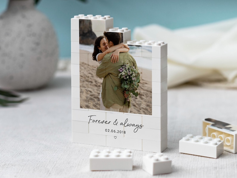 A white photo frame with a photo of a couple embracing, placed on a table with a vase and some Lego bricks.