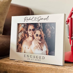 May include: A white rectangular picture frame featuring the black printed text Rachel & Samuel at the top and ENGAGED 29.07.2022 at the bottom. The frame holds a photograph of a couple wearing sunglasses against a rocky background.