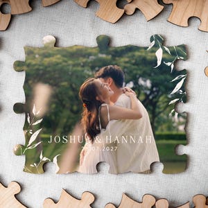 Wedding reception table setup featuring the personalised UV-printed wooden photo puzzle guest book alternative with a photograph of a couple embracing outdoors framed by white botanical leaves alongside pens and the matching wooden instruction stand.