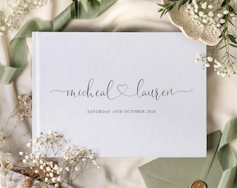 May include: A white rectangular guest book featuring the names micheal and lauren in elegant black cursive script with a small heart icon between them, followed by the text SATURDAY 14TH OCTOBER 2026 printed in a clean, simple black font.