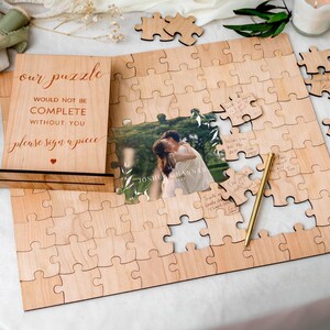 Overhead view of the assembled UV-printed wooden photo wedding jigsaw guestbook featuring a photograph of a couple embracing outdoors framed by white botanical leaves and text reading JOSHUA & HANNAH, and 28.07.2027, ready for guests to write their