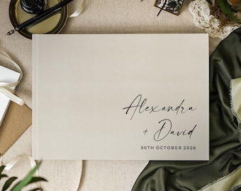 May include: A rectangular guest book with a light beige cover featuring the black printed text Alexandra + David and the date 30TH OCTOBER 2026. The book is positioned on a neutral surface alongside a black ink pen and a small framed photograph.