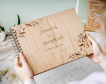 May include: A light-colored wooden guest book with a spiral binding. The cover features engraved botanical leaf designs on the corners and the text JOSHUA and HANNAH, OCTOBER 28 2025, and SOMERSET HOUSE, YORKSHIRE, centered on the front panel.
