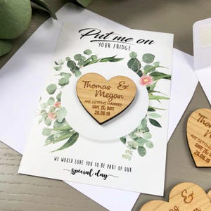 May include: A heart-shaped wooden magnet featuring the engraved text Thomas & Megan ARE GETTING MARRIED SAVE THE DATE 26.08.19, attached to a white card with green eucalyptus leaf illustrations and the text Put me on YOUR FRIDGE.