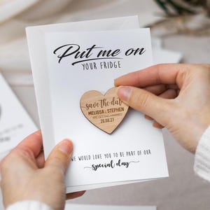 Op de afbeelding: Een witte rechthoekige kaart met de zwarte tekst Put me on YOUR FRIDGE en We would love you to be part of our special day. Een houten hartvormige magneet is in het midden bevestigd met de tekst save the date MELISSA & STEPHEN ARE GETTING MARRIED 26.08.27.