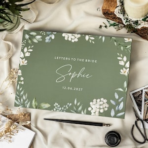 May include: A sage green guest book with the text "Letters to the Bride" and the name "Sophie" in elegant script. Floral illustrations of white flowers and green leaves frame the text. The date "12.06.2027" is also displayed.
