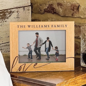 Può includere: Una cornice portafoto rettangolare in legno chiaro con il testo inciso THE WILLIAMS FAMILY in alto e la parola love in corsivo in basso a sinistra. La cornice contiene una fotografia di una famiglia di quattro persone che cammina all'aperto.