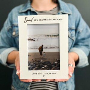 May include: A white rectangular picture frame featuring the black printed text Dad YOU ARE ONE IN A MILLION at the top and LOVE YOU LOTS, OLIVIA with a small heart symbol at the bottom, containing a photograph of a person and a child on a beach.