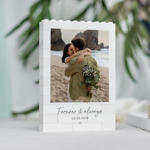 A white photo frame with a photo of a couple embracing on a beach, surrounded by greenery.