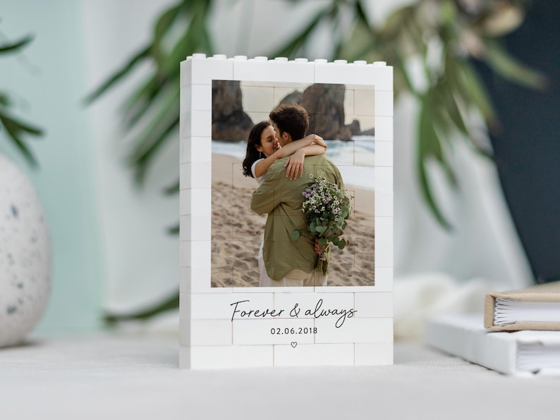 A white photo frame with a photo of a couple embracing on a beach, surrounded by greenery.