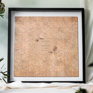 May include: A framed wooden puzzle with a black frame and white mat. The puzzle pieces are light brown and covered in handwritten messages. The center of the puzzle has the names "Joshua and Hannah" and the date "July 28 2023".