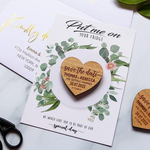 May include: A heart-shaped wooden magnet featuring the text "save the date THOMAS & REBECCA ARE GETTING MARRIED 30.07.2022 in Cyprus" attached to a white card with green eucalyptus leaf illustrations and the text "Put me on YOUR FRIDGE".