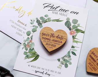 May include: A heart-shaped wooden magnet featuring the text "save the date THOMAS & REBECCA ARE GETTING MARRIED 30.07.2022 in Cyprus" attached to a white card with green eucalyptus leaf illustrations and the text "Put me on YOUR FRIDGE".