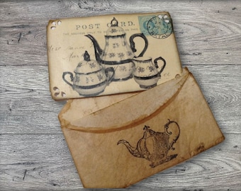 May include: A vintage postcard with a floral design of a teapot and two teacups. The postcard is folded in half and has a brown envelope with a black and white illustration of a teapot on the front. The postcard says "Post Card. The address only to be written on this side."