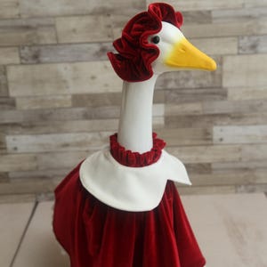 Pode incluir: Uma figura decorativa de ganso vestida com um vestido de veludo vermelho, um gorro a condizer e uma gola branca. O ganso tem um corpo branco, um bico amarelo e está posicionado contra um fundo de madeira clara.
