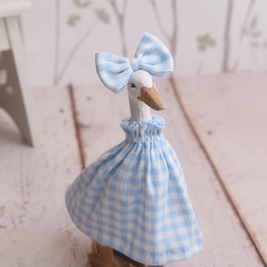 May include: A wooden goose figurine wearing a blue and white gingham dress and matching bow. The goose has a white head, a brown beak, and brown feet. The figurine is on a light wood surface.