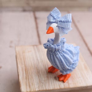 May include: A whimsical goose figurine, adorned in a light blue dress and matching bow. The goose has an orange beak and feet, standing on a small wooden block. The figurine is a charming decorative item.