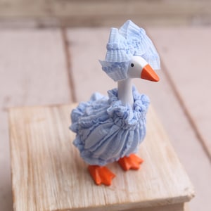 May include: A whimsical figurine of a goose, adorned in a light blue ruffled outfit and matching bonnet. The goose has a white head, an orange beak, and orange feet. It is posed on a small wooden block.
