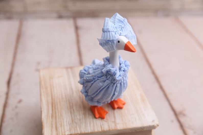 May include: A whimsical figurine of a goose, adorned in a light blue ruffled outfit and matching bonnet. The goose has a white head, an orange beak, and orange feet. It is posed on a small wooden block.