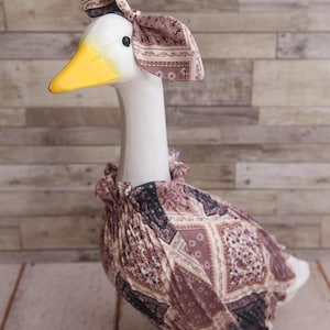 May include: A ceramic goose figurine with a white body, yellow beak, and a patterned headscarf and dress in shades of purple, brown, and cream. The goose is perched on a green base.