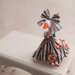 May include: A white goose figurine wearing a fabric dress and a matching bow on its head. The dress and bow feature a pattern of vertical green stripes and small brown teddy bear illustrations inside red oval frames. The goose has an orange beak.