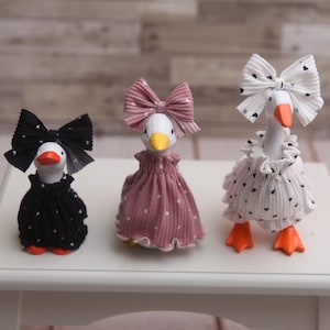 Op de afbeelding: Drie decoratieve ganzenbeeldjes met witte lichamen, oranje snavels en poten, gekleed in verschillende outfits. Eén gans draagt een zwarte jurk met witte stippen en een bijpassende strik. Een andere draagt een roze jurk en strik. De derde draagt een witte jurk met zwarte hartjes en een bijpassende strik.