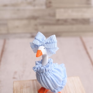May include: A whimsical goose figurine, adorned in a light blue ruffled dress and matching bow. The goose has an orange beak and feet, standing on a wooden surface. The background is a light-colored, textured wall.