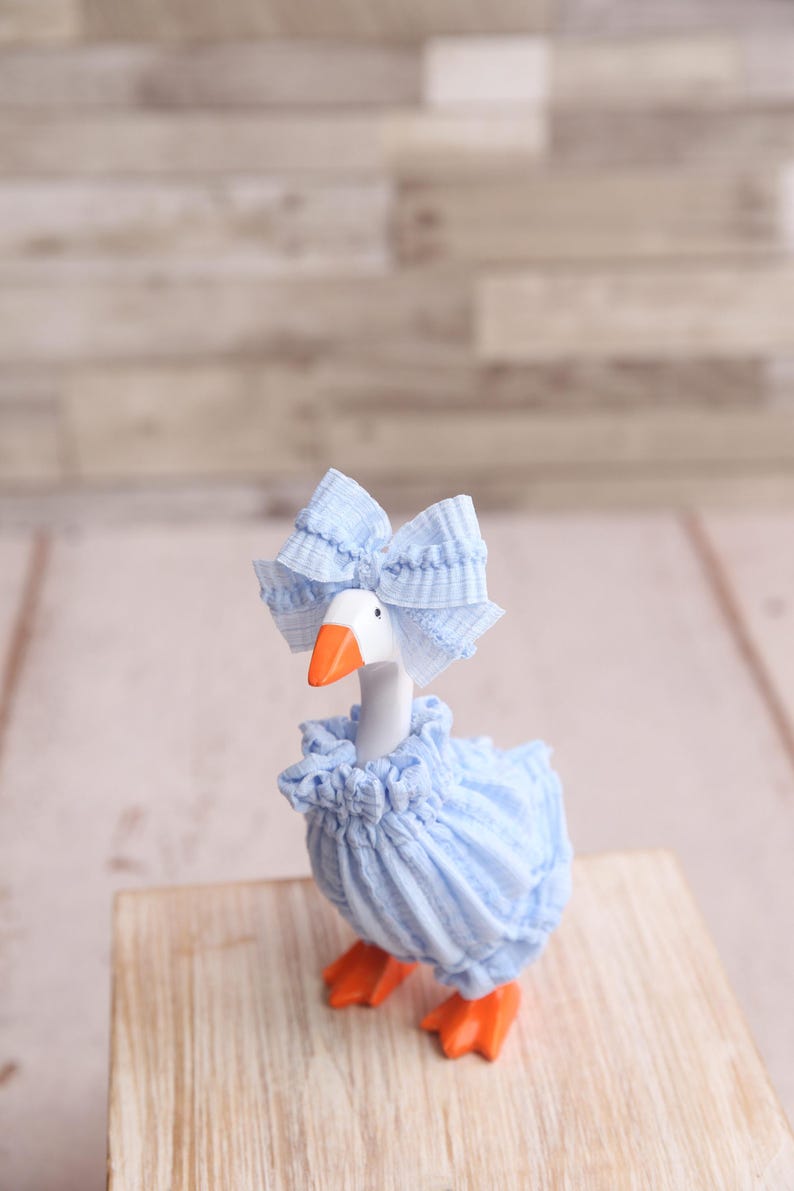 May include: A whimsical goose figurine, adorned in a light blue ruffled dress and matching bow. The goose has an orange beak and feet, standing on a wooden surface. The background is a light-colored, textured wall.