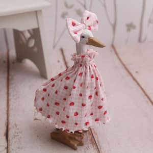 May include: A wooden goose figurine with a white head, a brown beak, and a pink dress adorned with red strawberries. The goose wears a matching bow on its head. The figurine is standing on a wooden base.