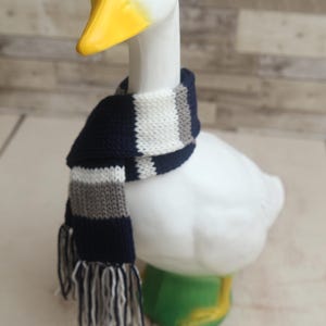 May include: A white ceramic goose figurine with a yellow beak and black eyes, wearing a navy, gray, and white striped scarf. The goose is perched on a green base.