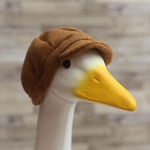 Peut inclure: Une tête de canard décorative avec une casquette marron. Le canard a un cou blanc et un bec jaune vif. La casquette est de couleur marron chaud, ajoutant une touche de caractère au design. L'arrière-plan est un motif de grain de bois clair.