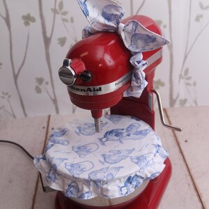 以下が含まれることがあります： 白と青の模様のカバーとリボンが付いた赤いKitchenAidスタンドミキサー。カバーとリボンは、白い背景に青い鳥の繰り返しデザインが特徴です。ミキサーは白い木の表面にあります。