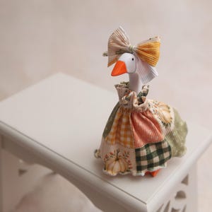 May include: A white goose figurine with an orange beak, wearing a fabric dress featuring a patchwork pattern of pumpkins, orange gingham, and green plaid. The goose also wears a matching fabric bow on its head, secured around its neck.