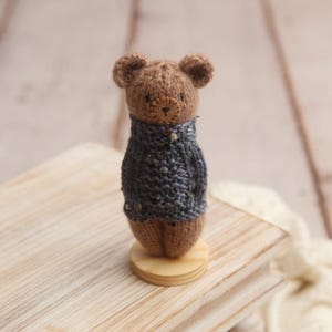 May include: A brown knitted bear wearing a blue knitted sweater stands on a wooden base. The bear has a small, round nose and black eyes.