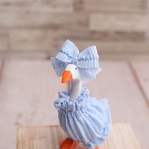 May include: A whimsical goose figurine with a white body, orange beak and feet, and a light blue ruffled dress and bow. The goose is posed on a wooden surface against a blurred background.