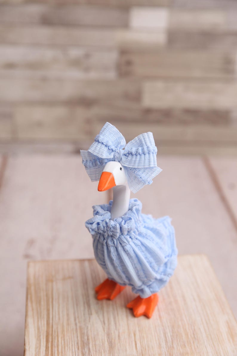 May include: A whimsical goose figurine with a white body, orange beak and feet, and a light blue ruffled dress and bow. The goose is posed on a wooden surface against a blurred background.
