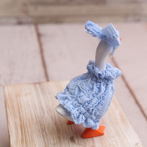 May include: A ceramic goose figurine wearing a light blue ruffled dress and matching bonnet. The goose has orange feet and a white body. The figurine is posed on a wooden block against a blurred background.