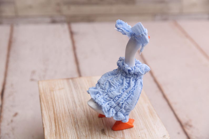 May include: A ceramic goose figurine wearing a light blue ruffled dress and matching bonnet. The goose has orange feet and a white body. The figurine is posed on a wooden block against a blurred background.
