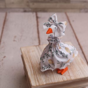 May include: A whimsical goose figurine, adorned with a white dress and matching bow, both patterned with green leaves and yellow fruit. The goose has an orange beak and feet. It is posed on a small wooden block, set against a light wood background.