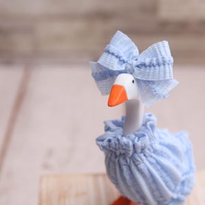 May include: A decorative goose figurine with a white head, orange beak, and blue bow. The goose wears a blue ruffled dress and a matching bonnet. The figurine is set against a blurred wooden background.