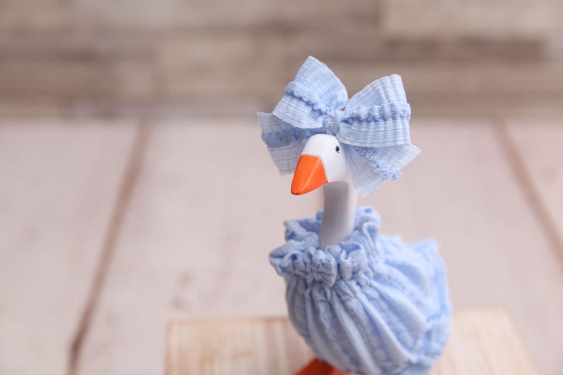 May include: A decorative goose figurine with a white head, orange beak, and blue bow. The goose wears a blue ruffled dress and a matching bonnet. The figurine is set against a blurred wooden background.