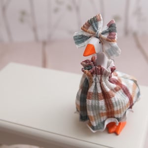 May include: A white goose figurine with an orange beak and feet, wearing a ruffled dress and a matching bow on its head. The fabric features a plaid pattern in muted shades of brown, green, and white. The goose stands upright on a surface.