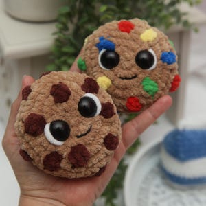 May include: Two plush, round, brown cookies with smiling faces and dark brown chocolate chips. One cookie has colorful candy-coated chocolate pieces. The cookies have black eyes and white highlights. The cookies are handmade and appear soft.