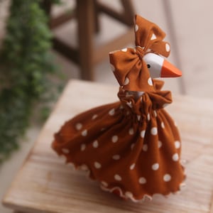 Könnte beinhalten: Eine skurrile Gänsefigur mit weißem Kopf, orangefarbenem Schnabel und einem braunen Kleid und Schleife mit weißen Punkten. Die Gans steht auf einer Holzoberfläche und wirkt rustikal. Die Figur ist ein dekoratives Objekt.