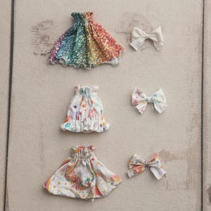 May include: Three doll dresses and two bows are displayed on a light wood surface. The dresses feature colorful patterns, including a rainbow, stars, and floral designs. The bows are white and patterned, complementing the dresses.