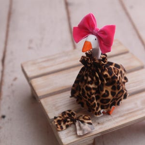 May include: A whimsical goose figurine with a bright pink bow on its head and a leopard print dress. A matching bow is placed near the goose on a wooden pallet. The goose has an orange beak and feet.