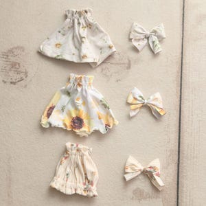 May include: Three small, ruffled skirts and three matching bows. The skirts feature floral patterns with yellow sunflowers and other flowers on a white background. The bows have a similar floral design.